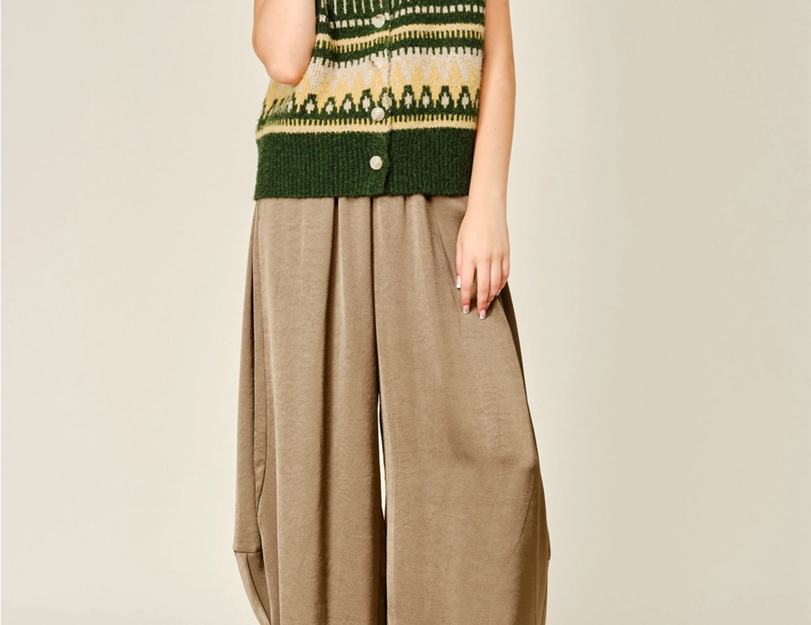 A model wearing a green and beige patterned knit vest with buttons, paired with loose brown wide-leg satin bloomer pants and brown suede mules, against a light beige studio background.