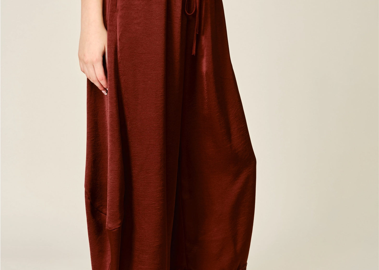 A person wearing loose, wide-leg rust-brown satin bloomer pants with a lace hem, styled with a gray sleeveless top and tan slip-on shoes against a plain light beige studio background.