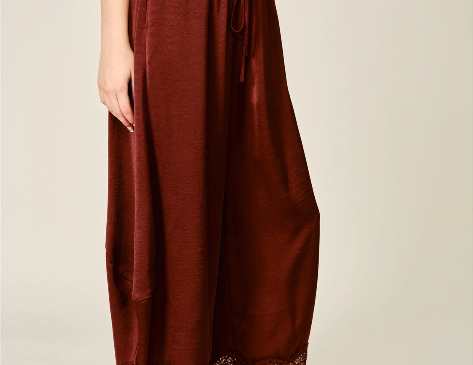 A person wearing loose, wide-leg rust-brown satin bloomer pants with a lace hem, styled with a gray sleeveless top and tan slip-on shoes against a plain light beige studio background.