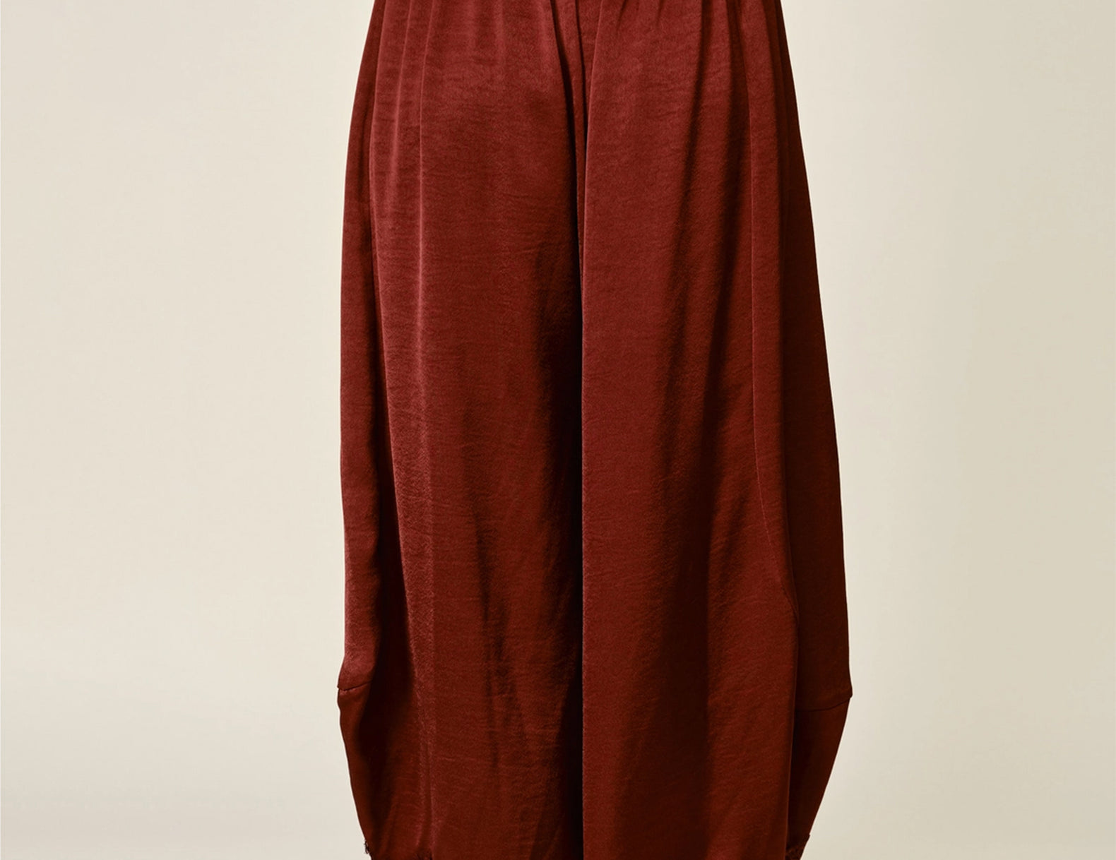 A person wearing loose, balloon-style bloomer pants in a deep rust red or burgundy fabric with a lace hem, paired with a gray top and tan slip-on slippers, photographed against a plain light beige studio background.