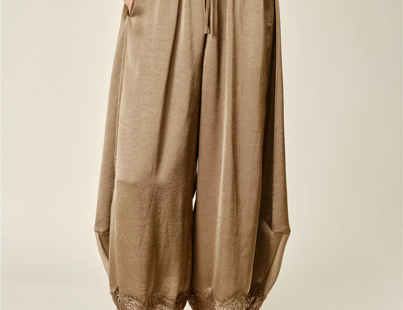 A person wearing loose, wide-leg taupe-brown satin bloomer pants with a drawstring waist and lace trim at the hems, paired with tan slip-on shoes against a plain light beige studio background.