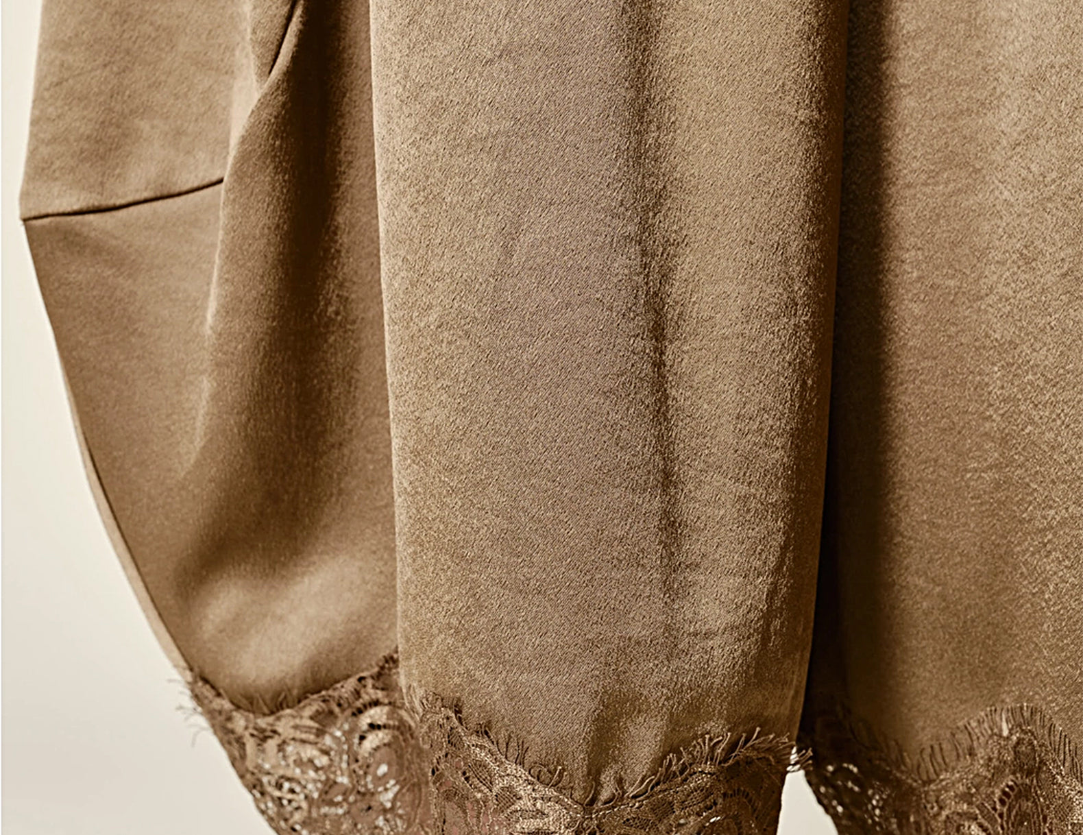 Close-up of a taupe-brown garment with voluminous draped fabric and a lace hem, shown against a plain light background.