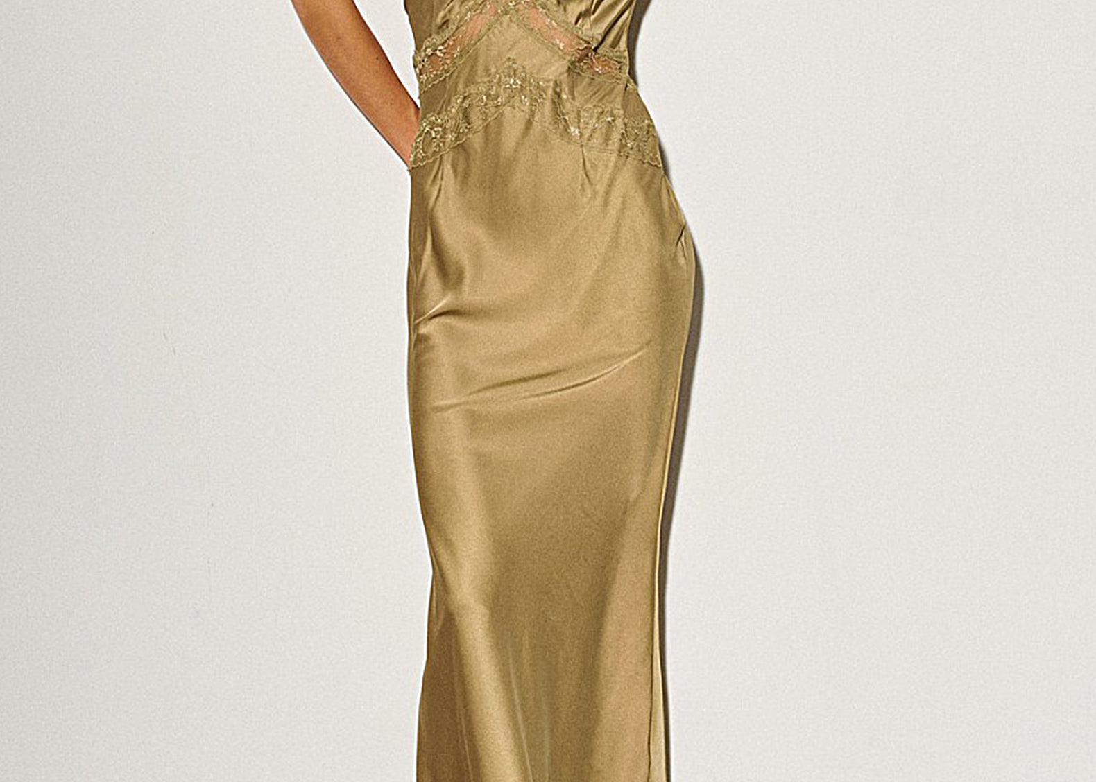 A woman models a floor-length satin slip gown in a warm metallic gold color with matching lace detailing along the neckline and waist. She is wearing black and white pointed-toe heels against a plain white wall.