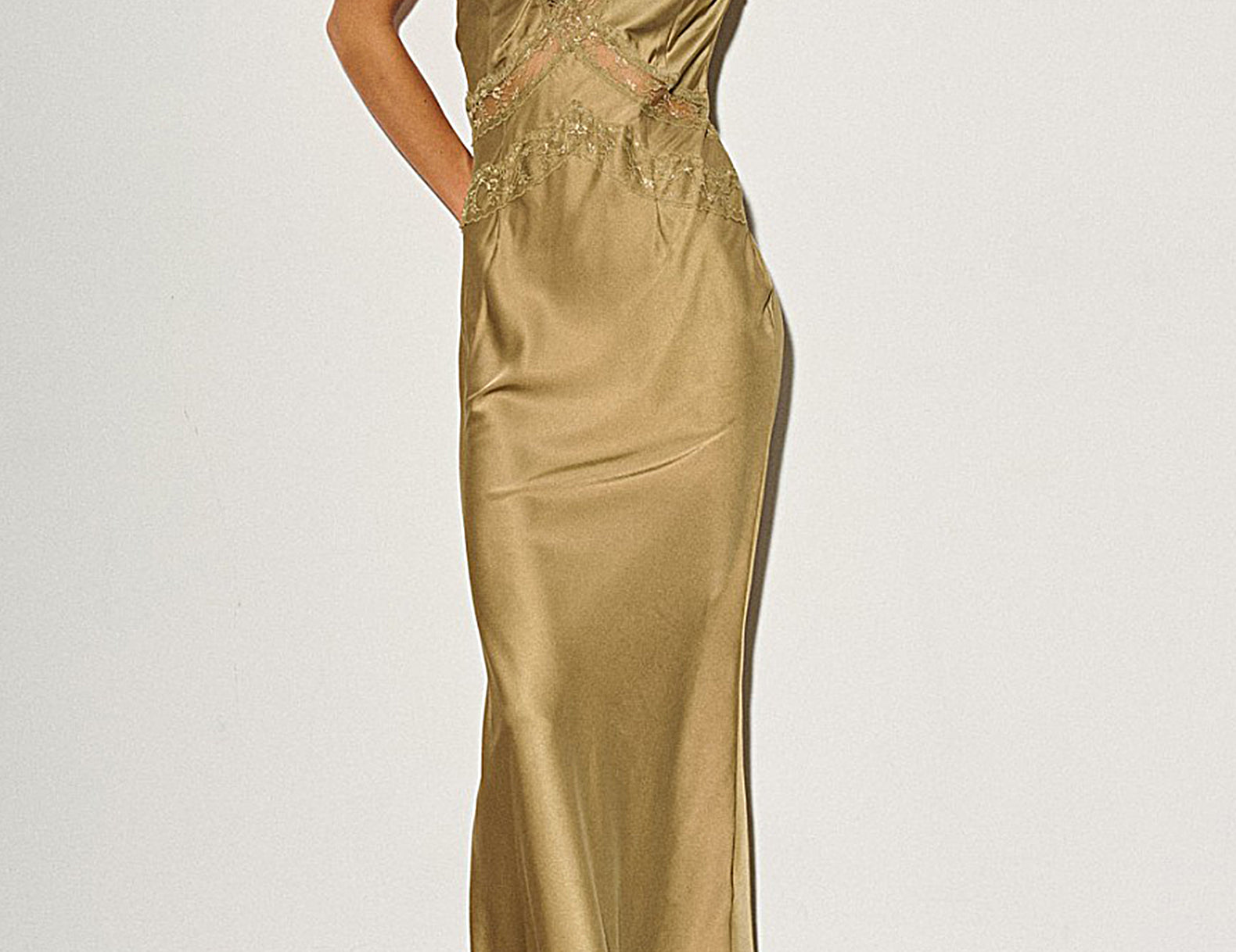 A woman models a floor-length satin slip gown in a warm metallic gold color with matching lace detailing along the neckline and waist. She is wearing black and white pointed-toe heels against a plain white wall.