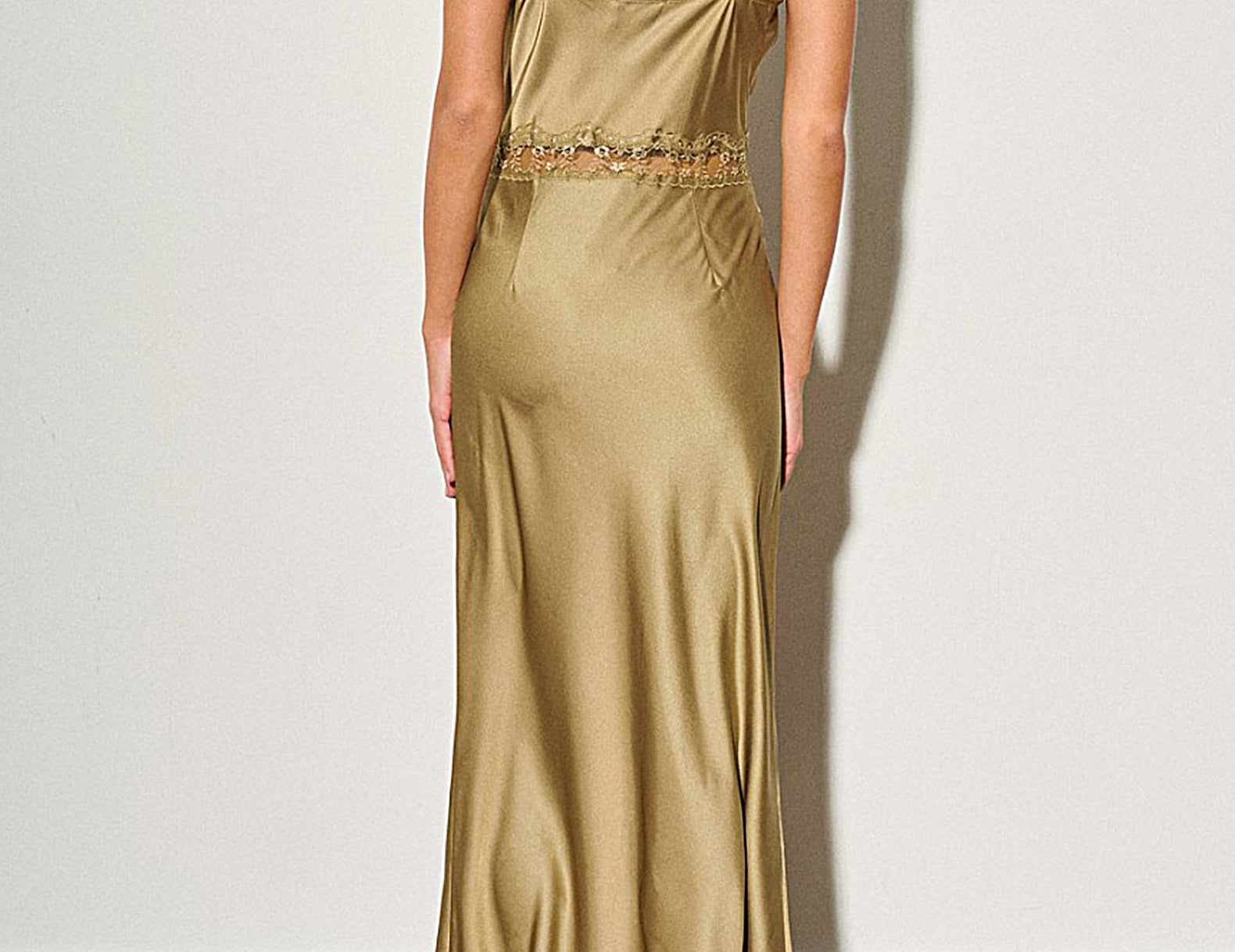 A woman viewed from behind wearing a floor-length satin slip dress in a warm metallic gold color, standing against a plain off-white wall.