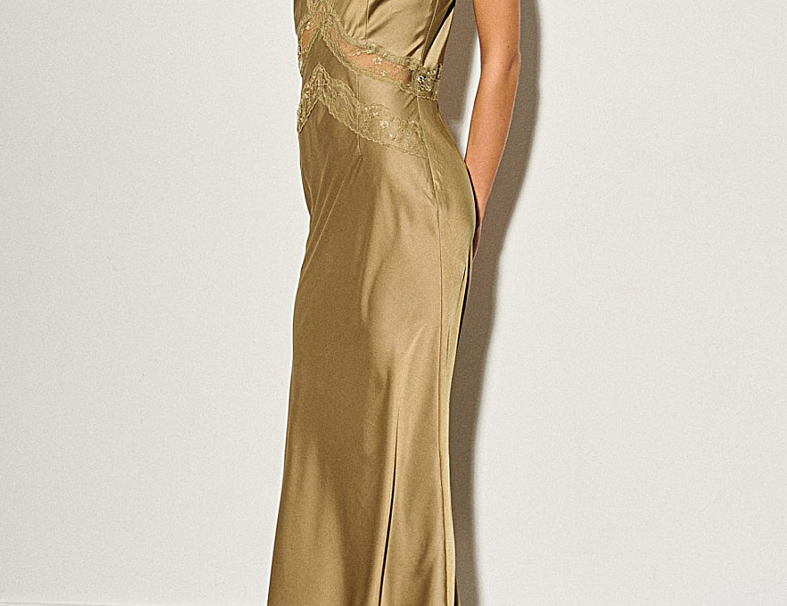 A woman models a floor-length champagne gold satin gown with a deep V-neckline, lace detailing at the bust, and side pockets, paired with black and beige pointed-toe flats against a plain off-white wall.