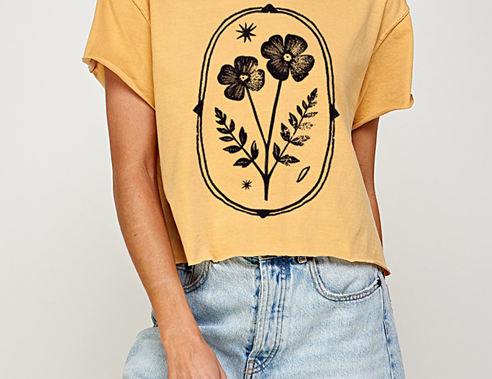 A woman wearing a cropped yellow t-shirt with a black floral graphic print and light-wash denim shorts, shown from the waist up against a light gray studio background.