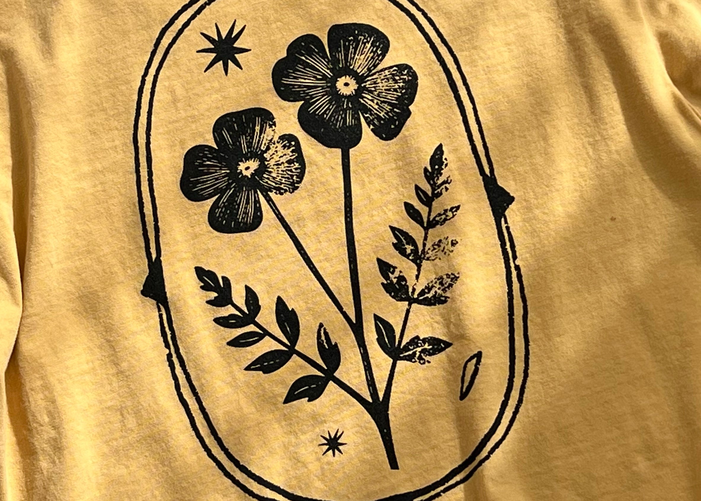 A close-up of a mustard yellow t-shirt featuring a black graphic print of two flowers, leaves, and stars inside an oval frame.