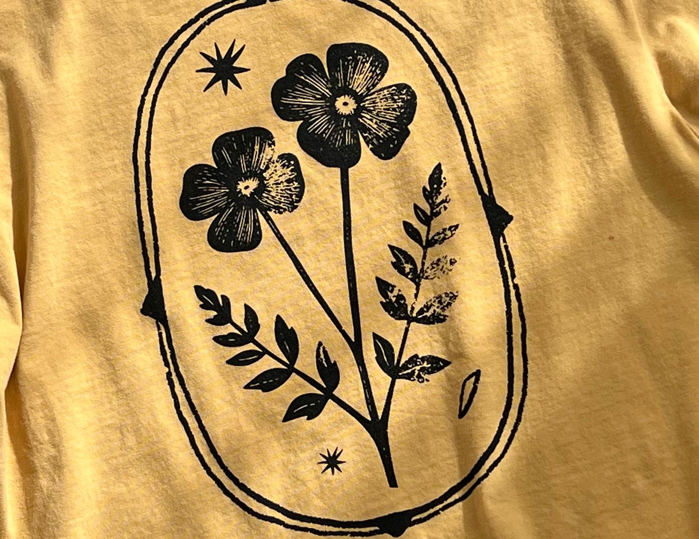 A close-up of a mustard yellow t-shirt featuring a black graphic print of two flowers, leaves, and stars inside an oval frame.