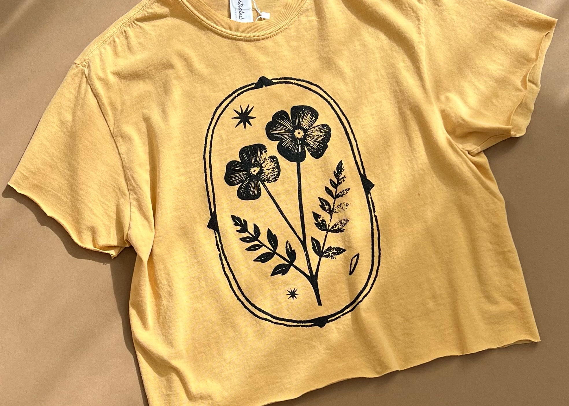 A pale yellow cropped t-shirt with a black floral graphic printed on the front, laid flat on a warm beige background with soft diagonal shadows.