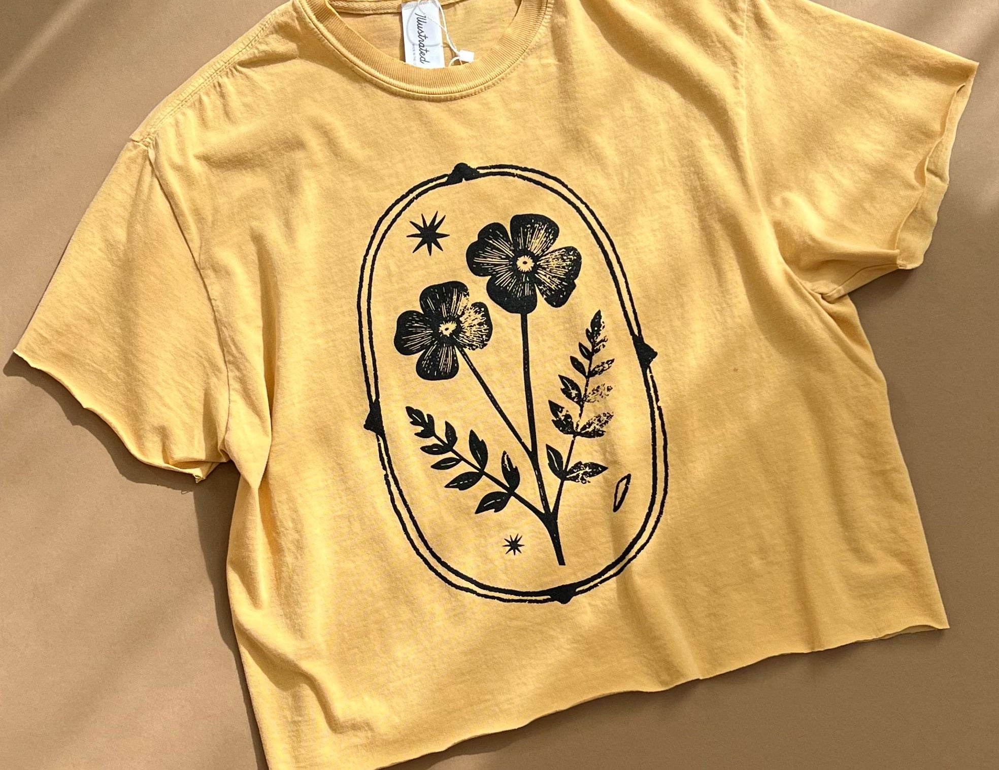 A pale yellow cropped t-shirt with a black floral graphic printed on the front, laid flat on a warm beige background with soft diagonal shadows.