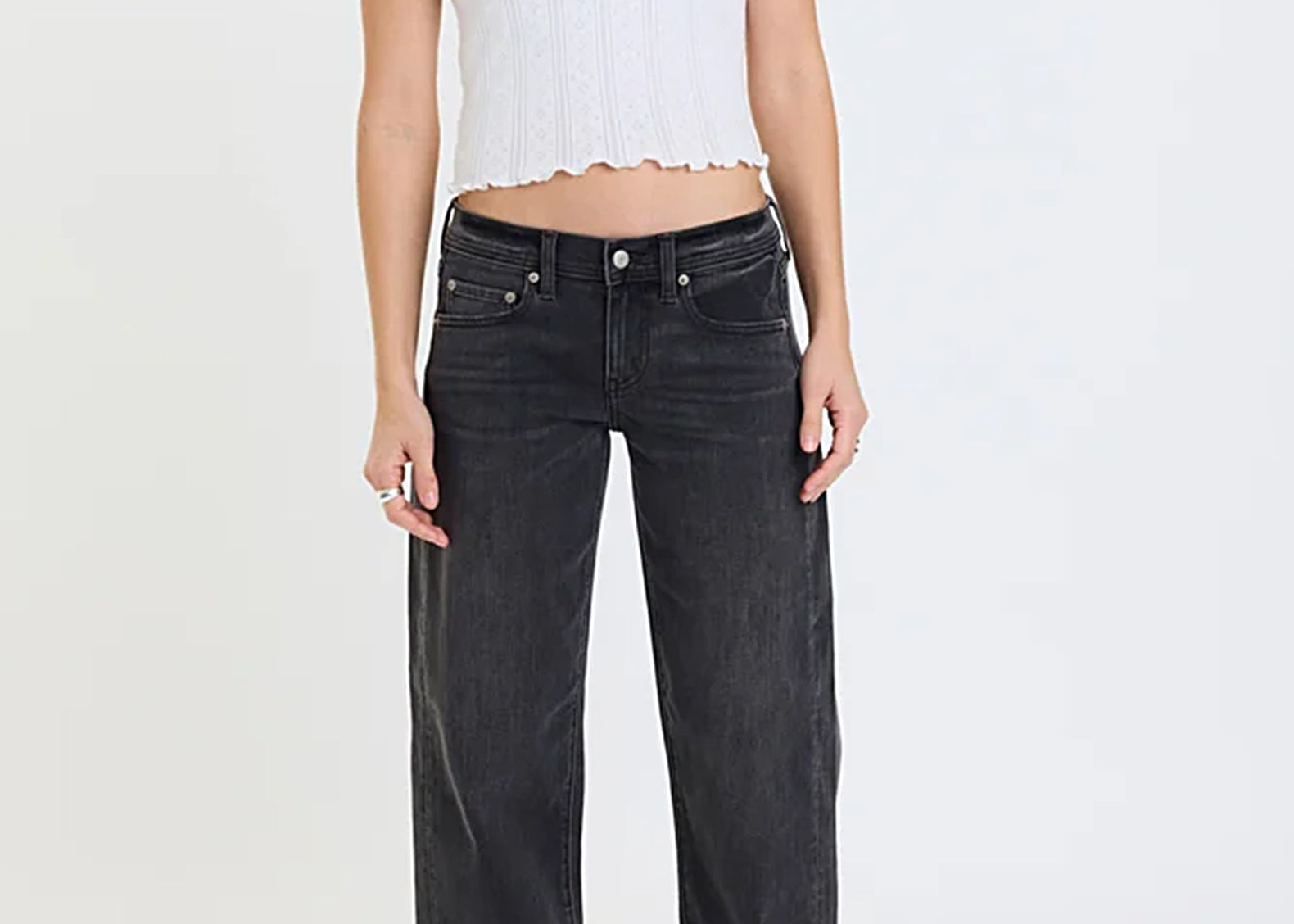 A model wearing a white short-sleeve ribbed knit crop top with scalloped edges and dark gray wide-leg jeans, standing against a plain white studio background.