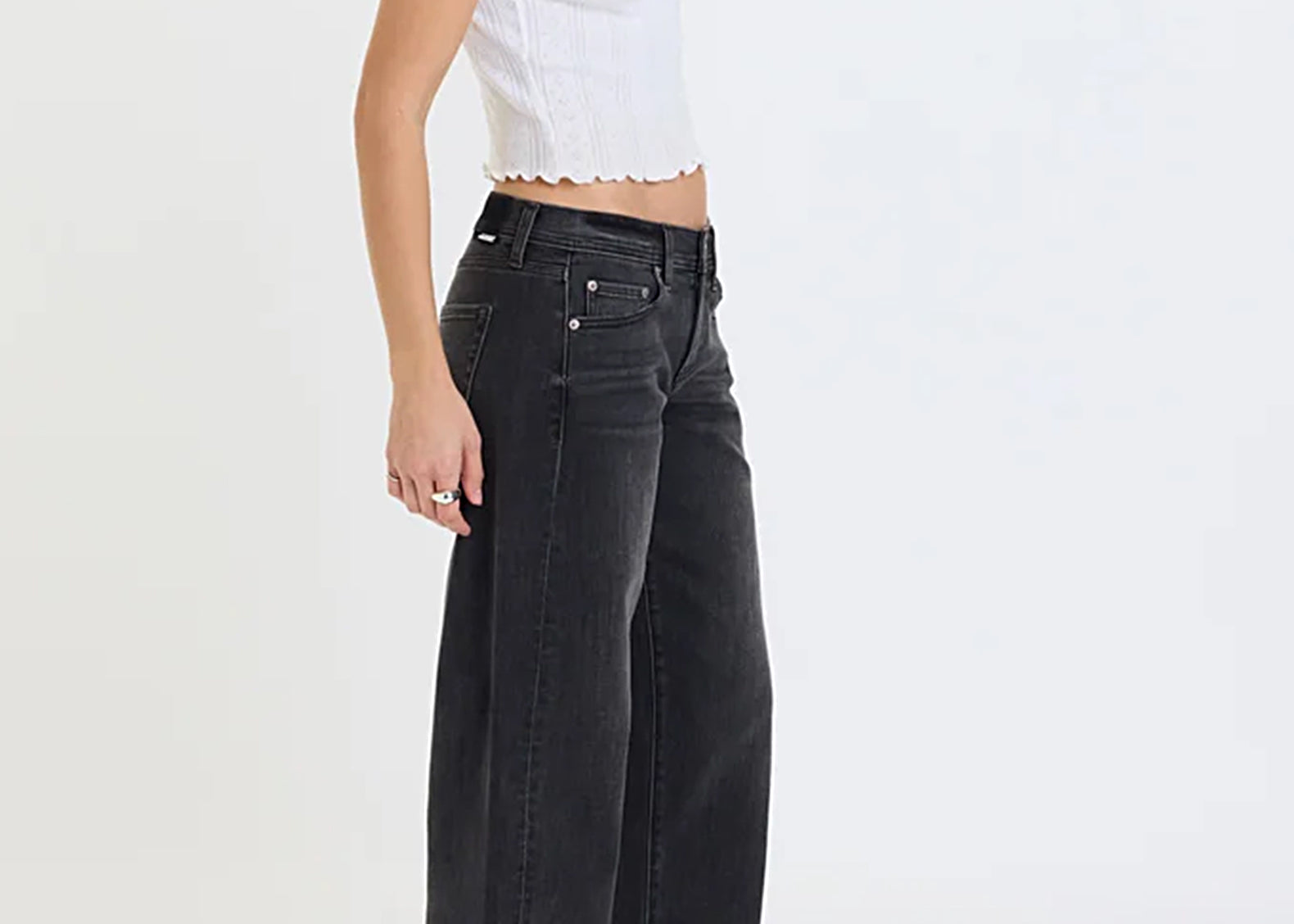 A model wearing black wide-leg denim jeans, a white cropped ribbed top, and black leather shoes, shown from a side profile against a plain white studio background.