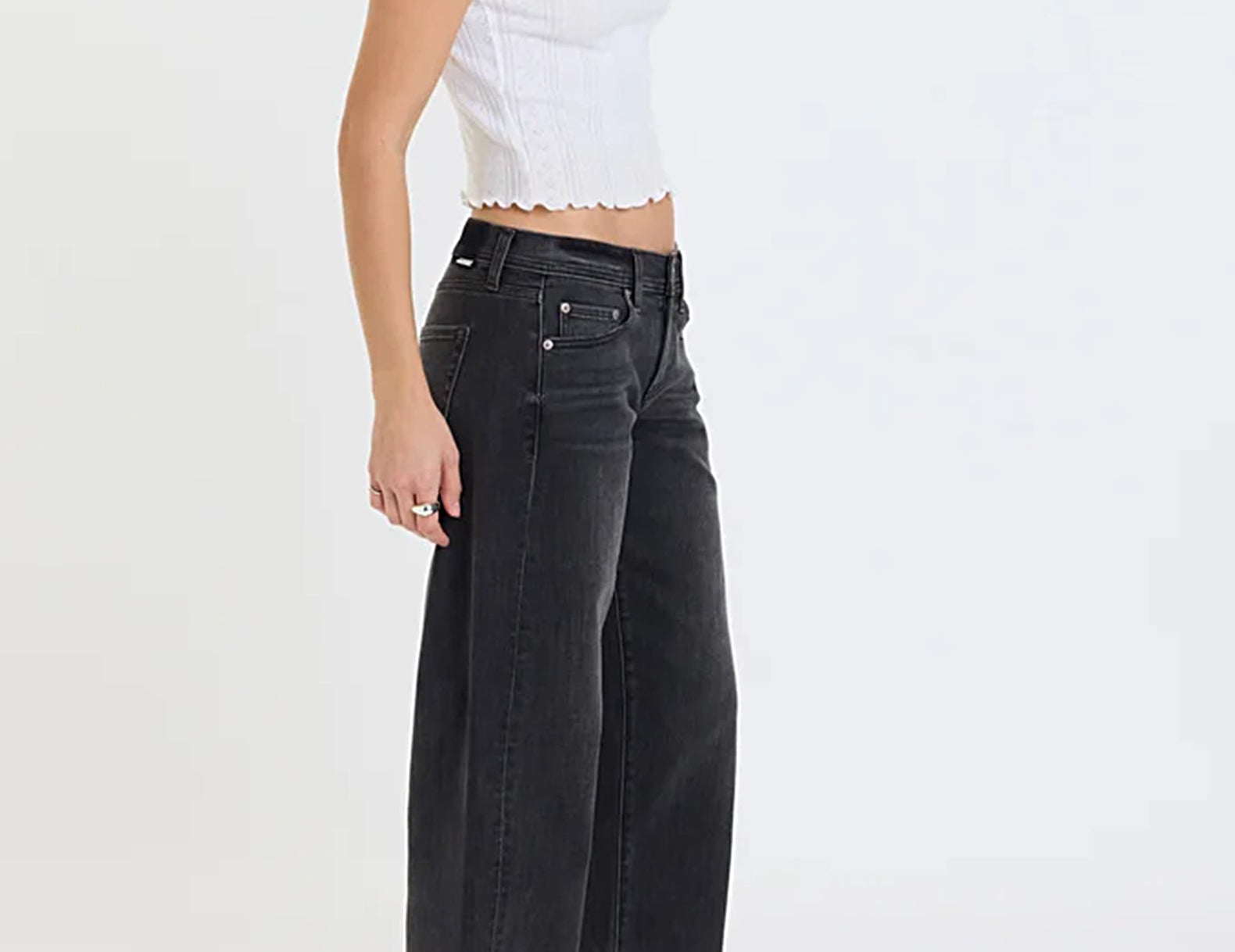 A model wearing black wide-leg denim jeans, a white cropped ribbed top, and black leather shoes, shown from a side profile against a plain white studio background.