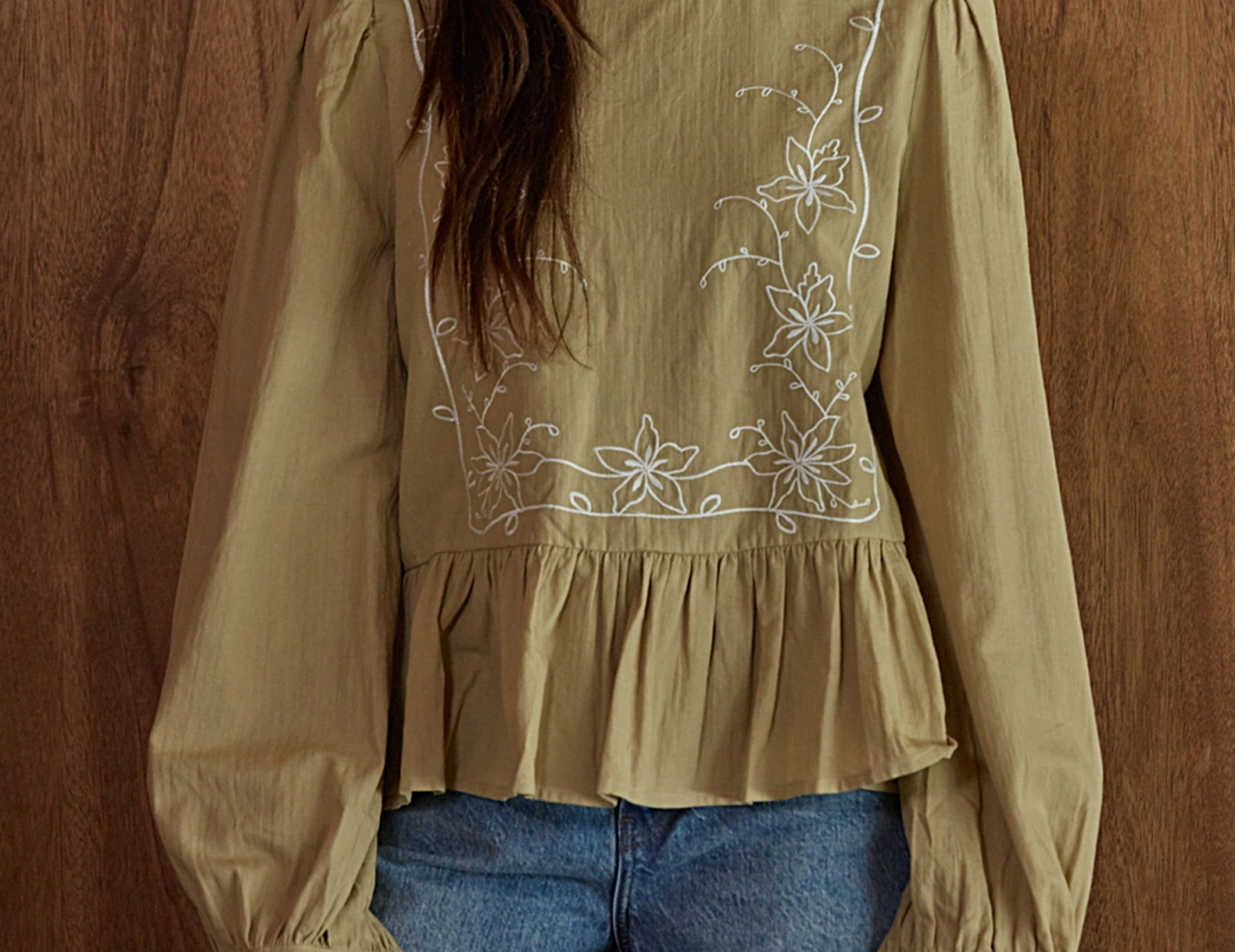 A woman wearing a beige long-sleeve blouse with white floral embroidery on the chest and cuffs, paired with blue jeans, standing against a wooden wall.