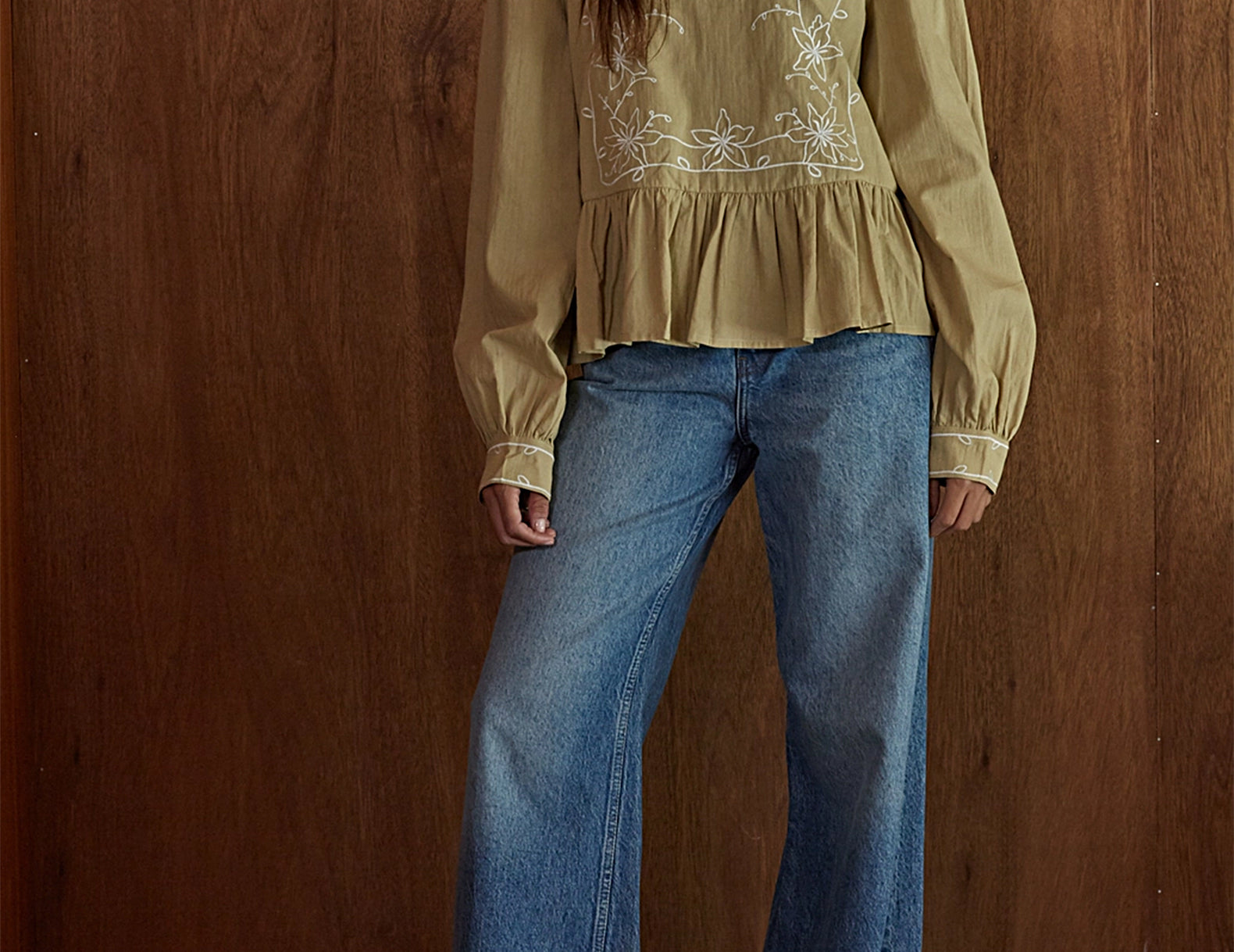 A woman wearing a beige long-sleeve blouse with white floral embroidery and blue wide-leg jeans, standing against a brown wooden wall.