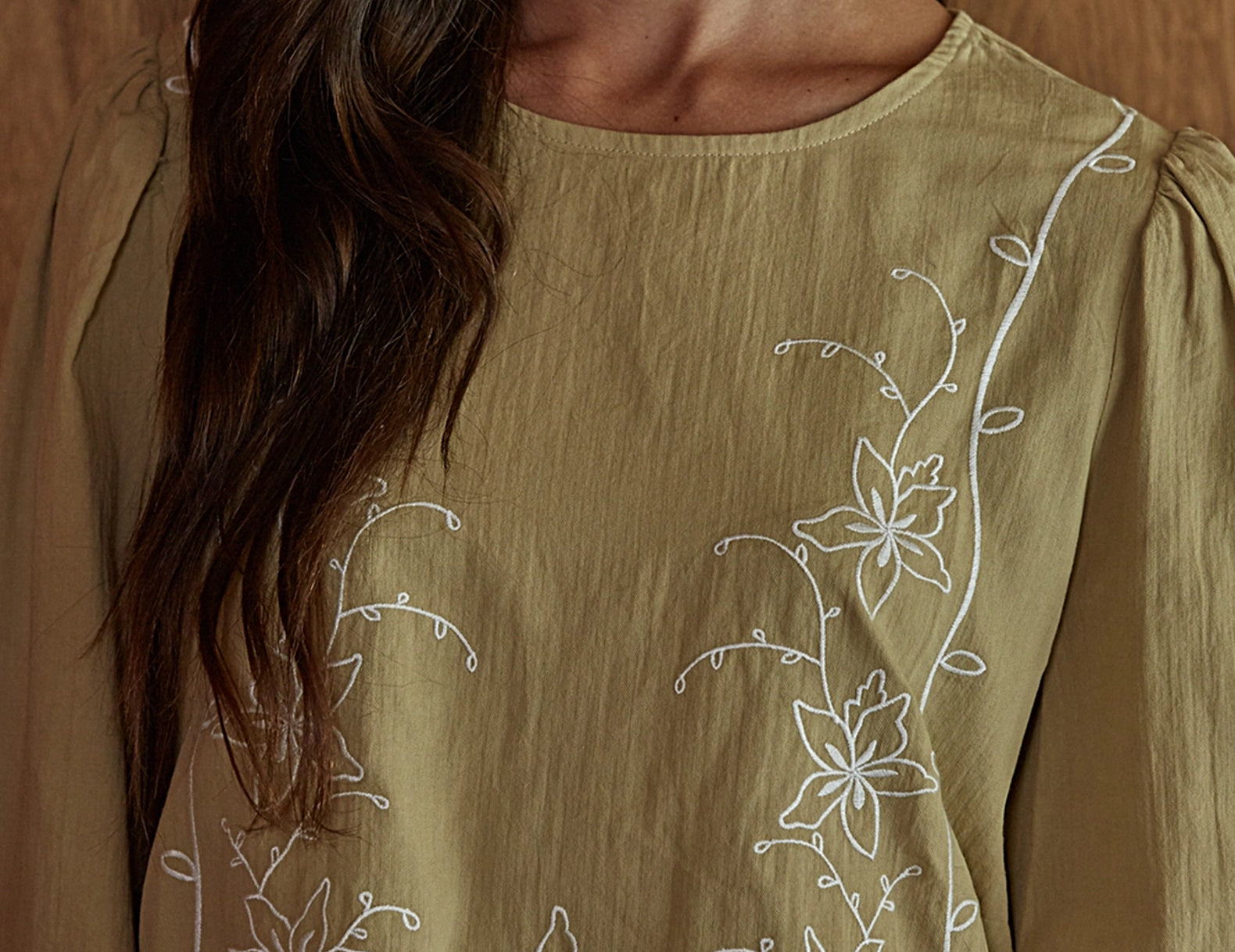 A close-up of a woman wearing a long-sleeve olive green top with white floral embroidery on the chest, set against a warm wooden background.