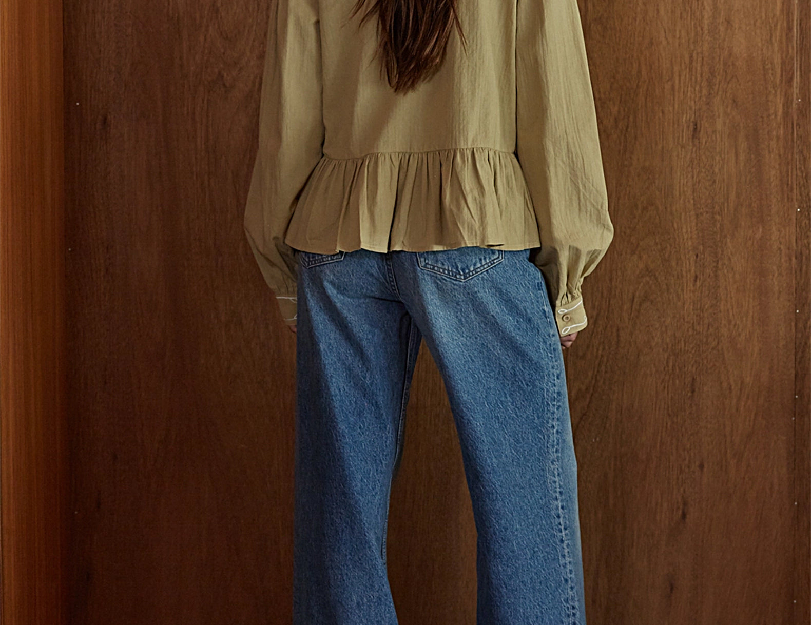 A woman viewed from behind wearing a beige peplum blouse and wide-leg blue jeans, standing against a brown wooden wall.