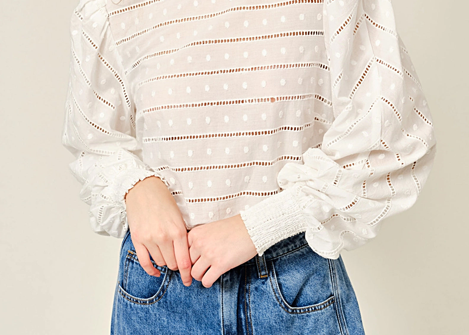 A woman wearing a white eyelet knit top with horizontal dotted lines and voluminous long sleeves, paired with blue jeans, against a plain light beige background.