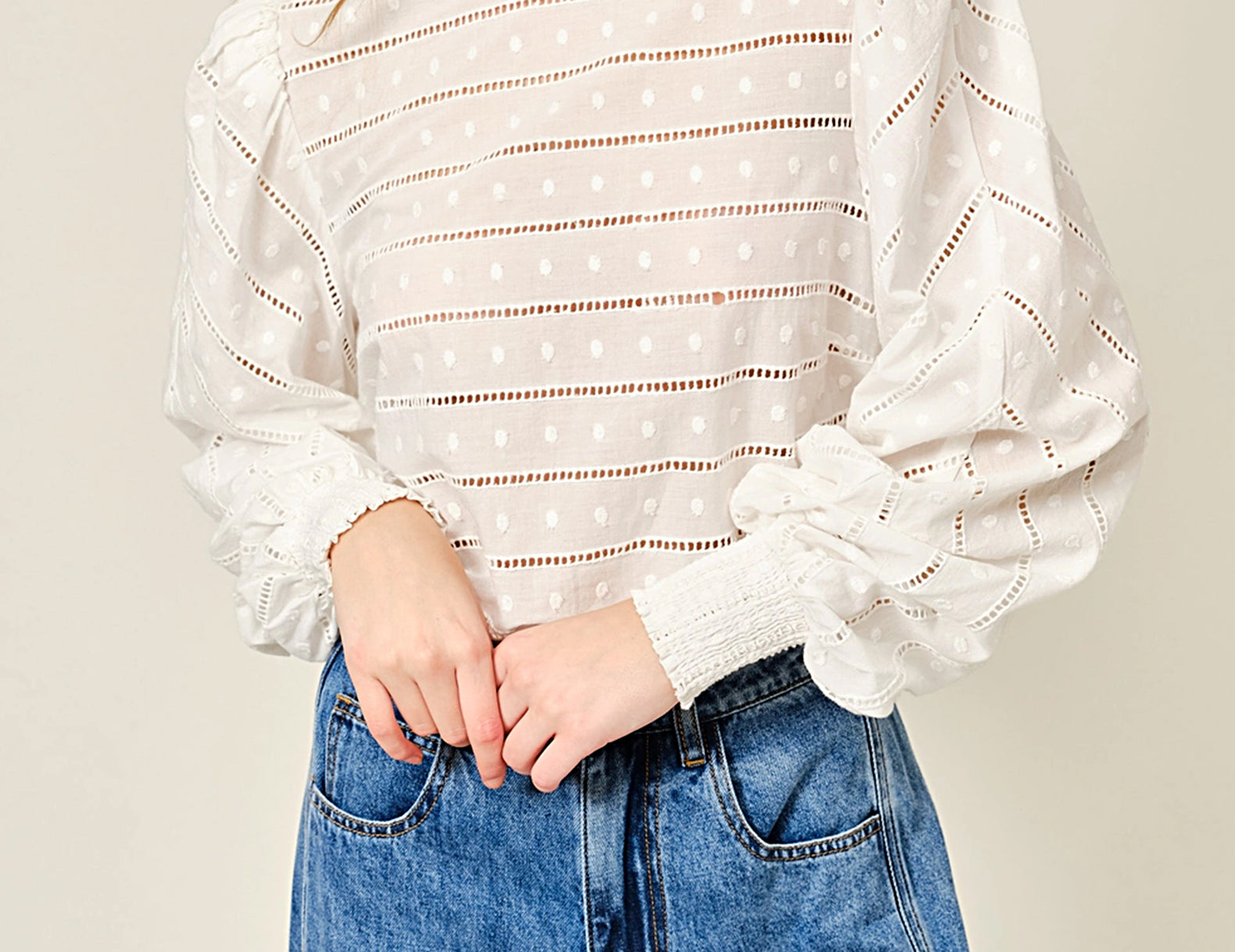 A woman wearing a white eyelet knit top with horizontal dotted lines and voluminous long sleeves, paired with blue jeans, against a plain light beige background.