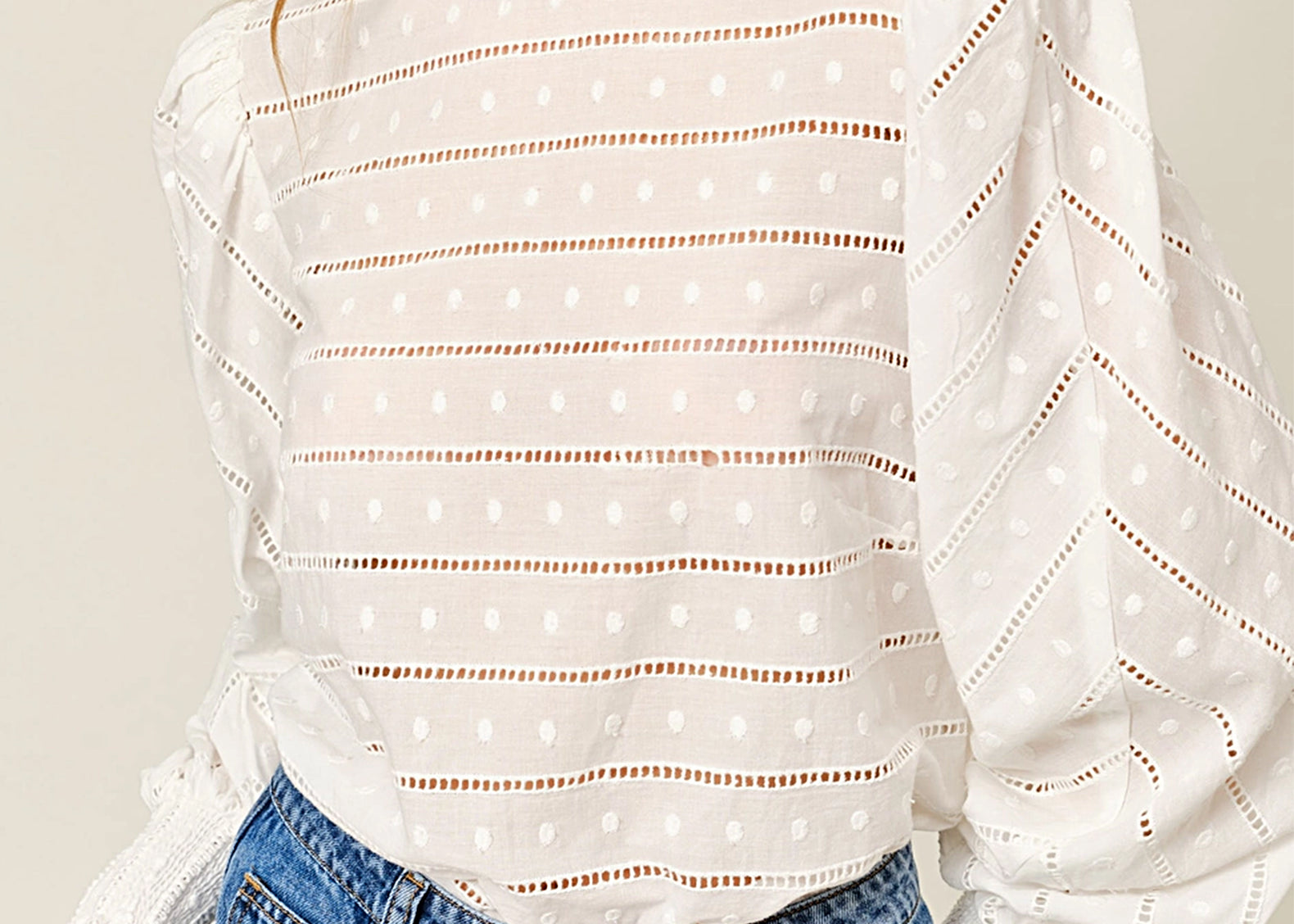 A woman wearing a white eyelet blouse with horizontal dotted cutout stripes and voluminous long sleeves, paired with blue jeans against a light beige studio background.