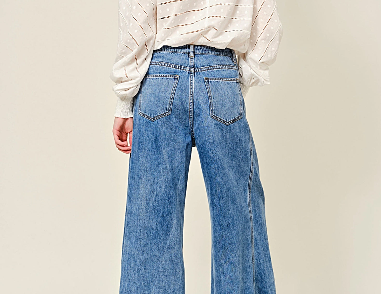 A woman shown from the back wearing a white textured long-sleeve top with a keyhole cutout and high-waisted, wide-leg blue denim jeans, paired with brown shoes, against a plain beige studio background.