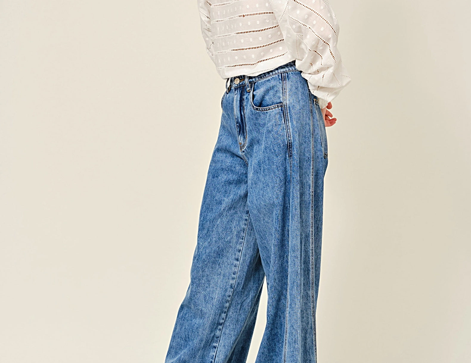 A model wearing a white textured long-sleeve blouse and high-waisted, wide-leg blue denim jeans with brown leather loafers, standing against a plain beige studio background.