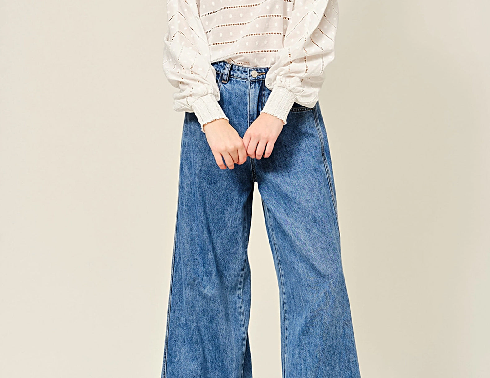 A model wearing a white long-sleeved blouse with sheer puff sleeves and wide-leg blue denim jeans, paired with brown leather loafers, against a plain beige studio background.