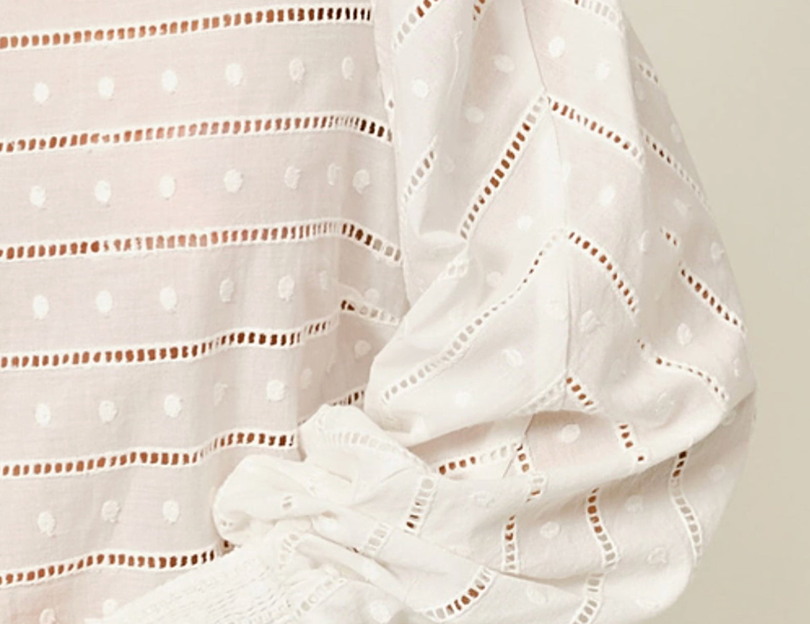 Close-up of a white eyelet blouse with horizontal dotted cutout stripes and gathered puff sleeves, worn with blue jeans against a light neutral background.