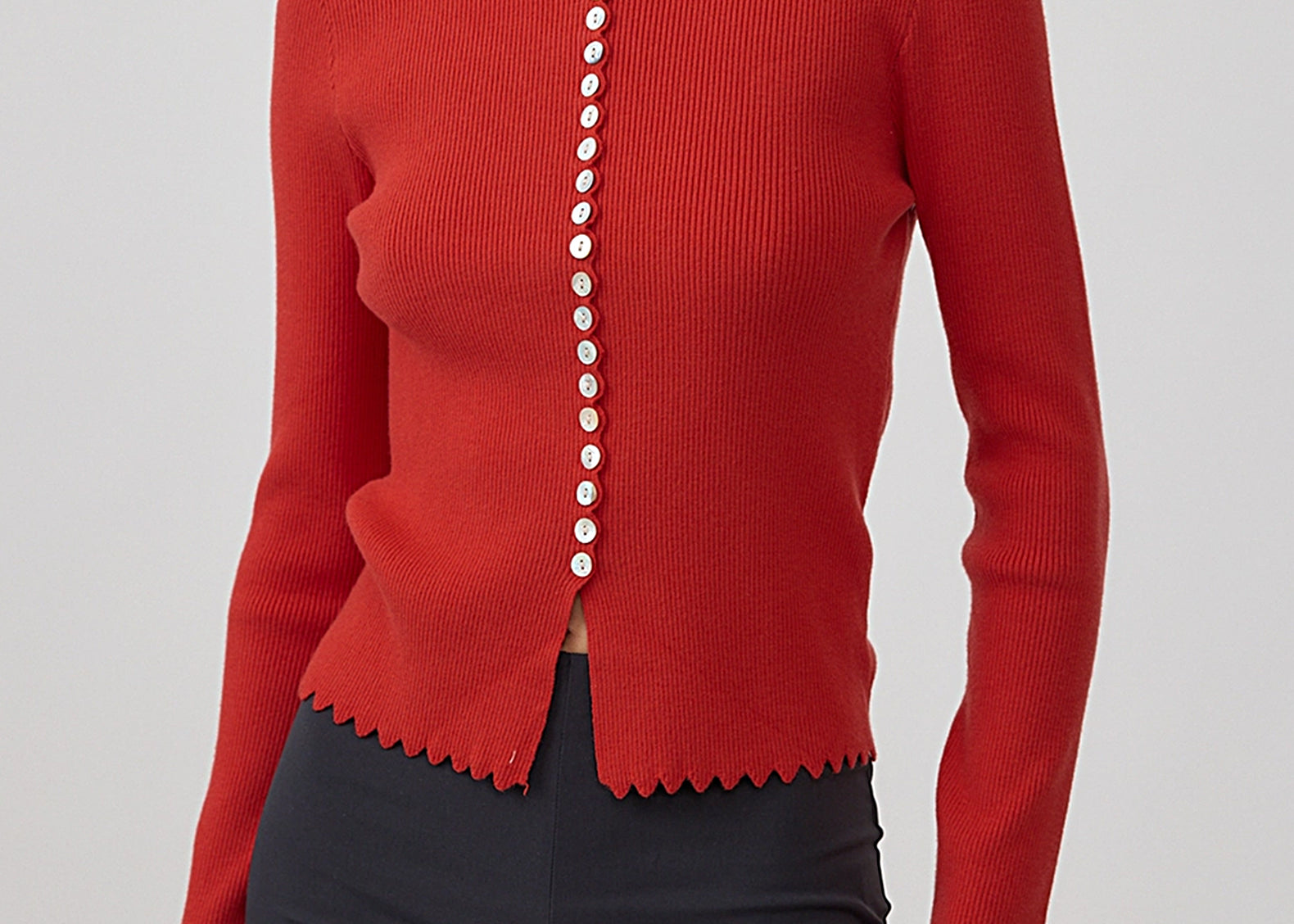 A woman wearing a bright red ribbed knit cardigan with scalloped edges and a vertical row of white buttons down the front, paired with dark charcoal pants, against a light gray studio background.