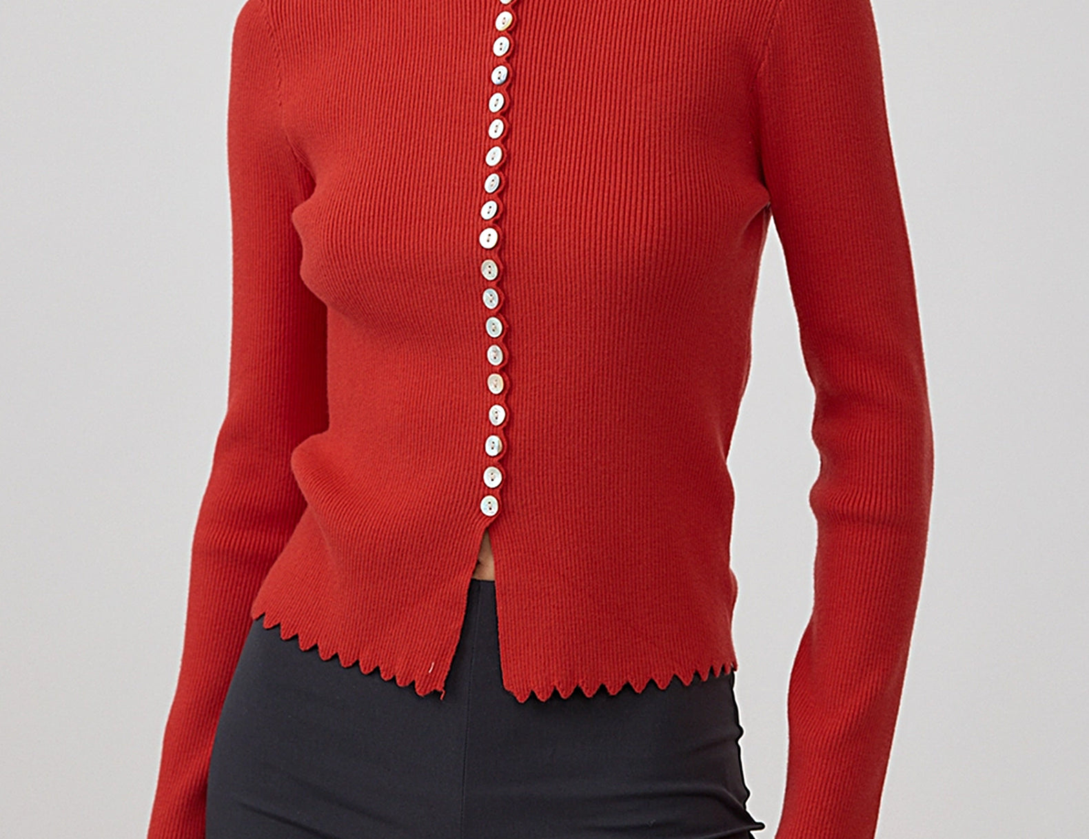 A woman wearing a bright red ribbed knit cardigan with scalloped edges and a vertical row of white buttons down the front, paired with dark charcoal pants, against a light gray studio background.