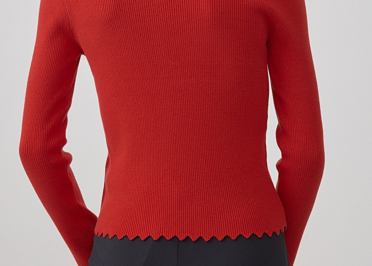 A woman shown from the back wearing a fitted ribbed knit sweater in a vivid red color, paired with dark charcoal pants, against a plain light gray studio background.