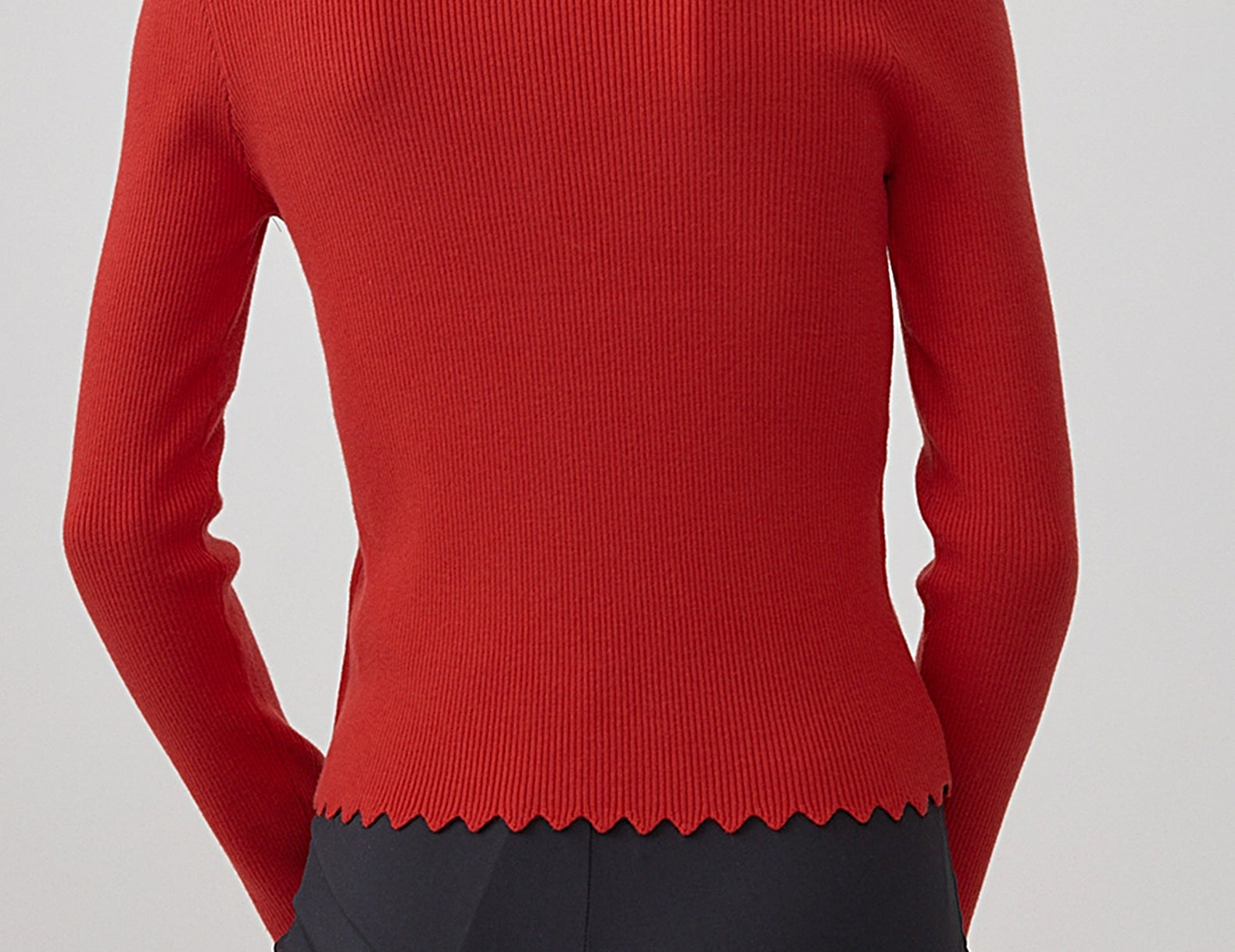 A woman shown from the back wearing a fitted ribbed knit sweater in a vivid red color, paired with dark charcoal pants, against a plain light gray studio background.