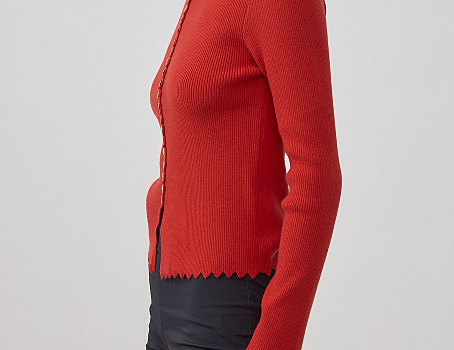 A woman wearing a red ribbed knit cardigan with scalloped edges and dark trousers, shown in a side profile against a light gray studio background.