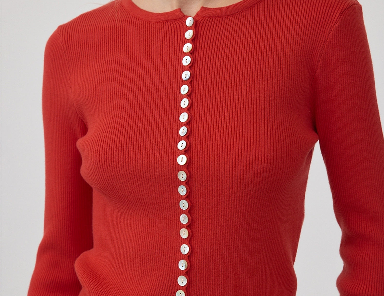 A close-up of a model wearing a vibrant red ribbed knit cardigan with a scalloped hem and a vertical row of white buttons down the front.