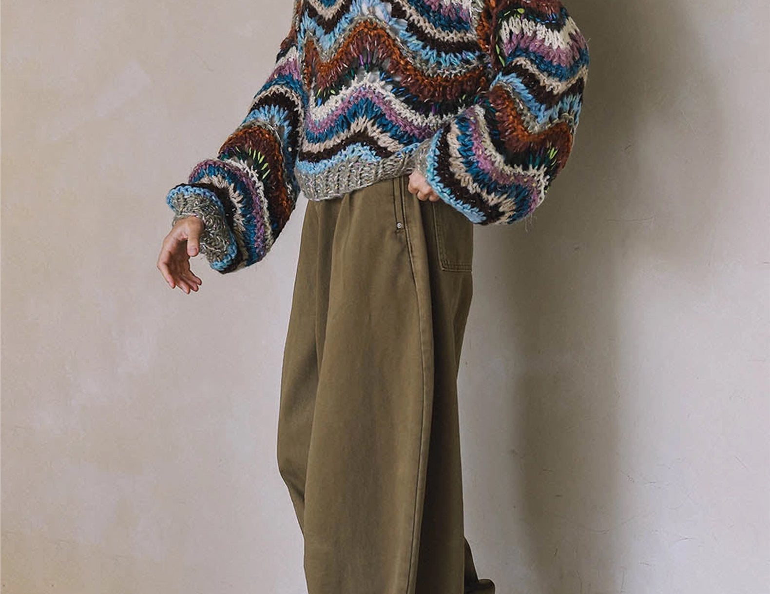 A person wearing a chunky, multicolor zigzag knit sweater with brown, blue, pink, and white stripes, paired with wide-leg olive green pants and white sneakers, standing against a textured beige wall.