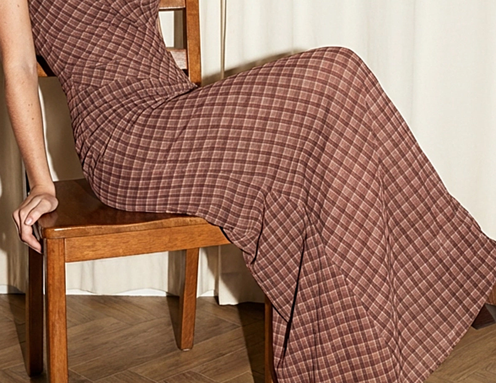 A woman seated on a wooden chair wearing a brown plaid maxi dress with thin straps and brown pointed-toe boots, in an indoor setting with a cream curtain and wood floor.