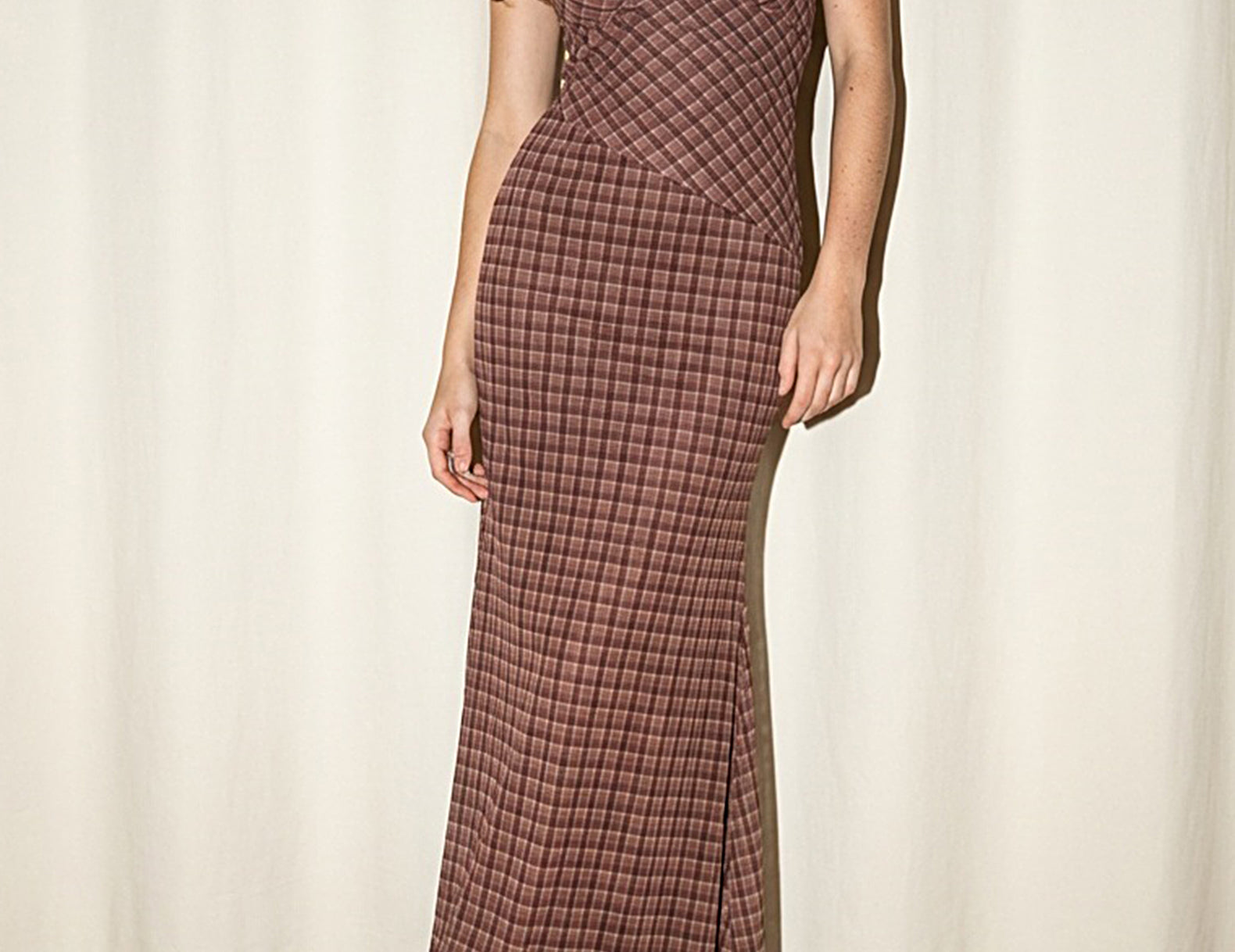 A woman models a brown and red plaid maxi dress with spaghetti straps and a wrap-style bodice, paired with brown suede boots, set against a cream-colored curtain backdrop.