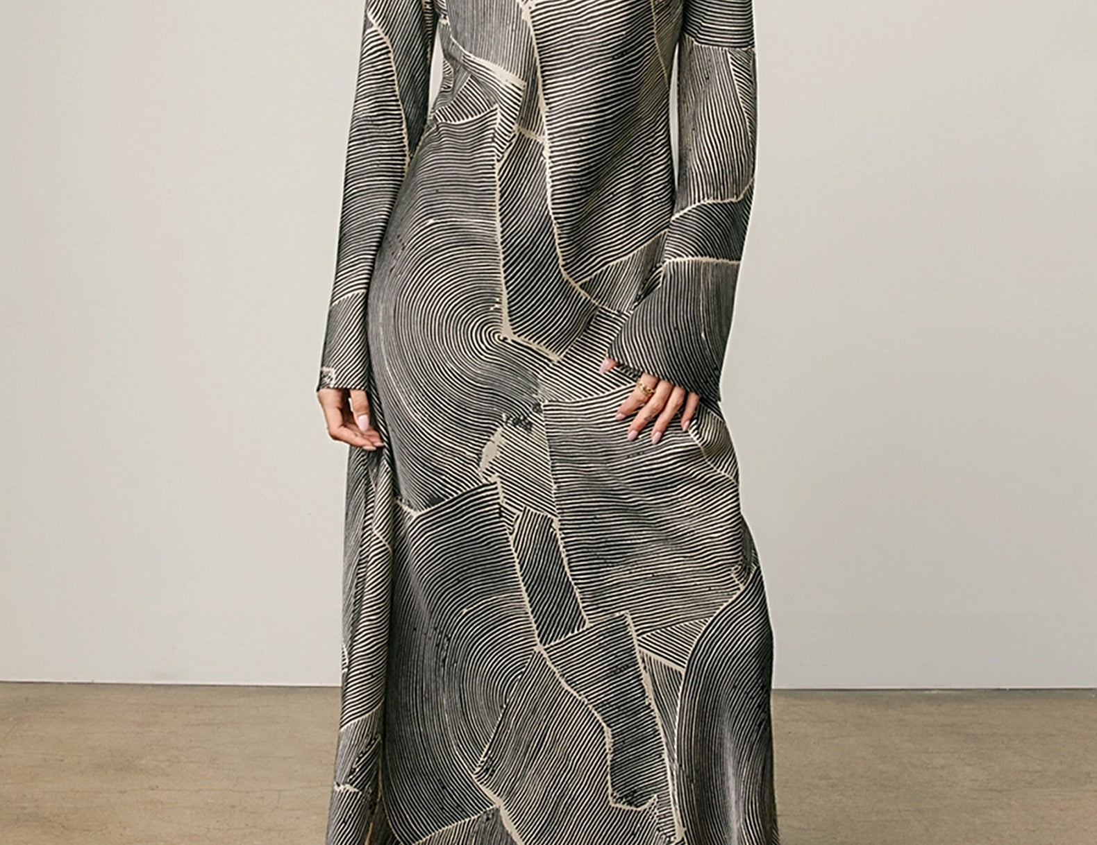 A woman modeling a long-sleeve maxi dress with a gray and black abstract wood-grain pattern and cream lines, paired with beige heeled sandals against a neutral studio backdrop.