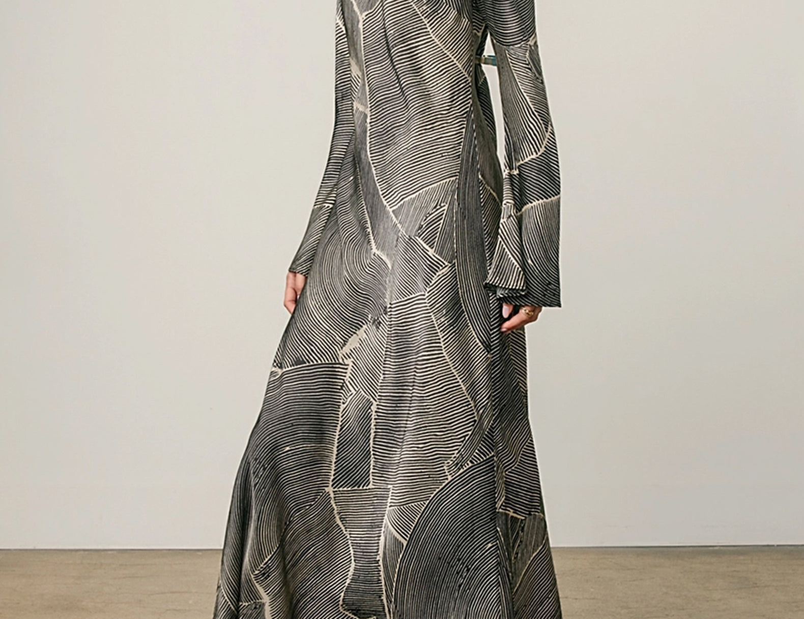 A woman models a long-sleeved maxi dress with a black, gray, and white abstract leaf-like pattern, paired with beige heeled sandals against a neutral studio backdrop.