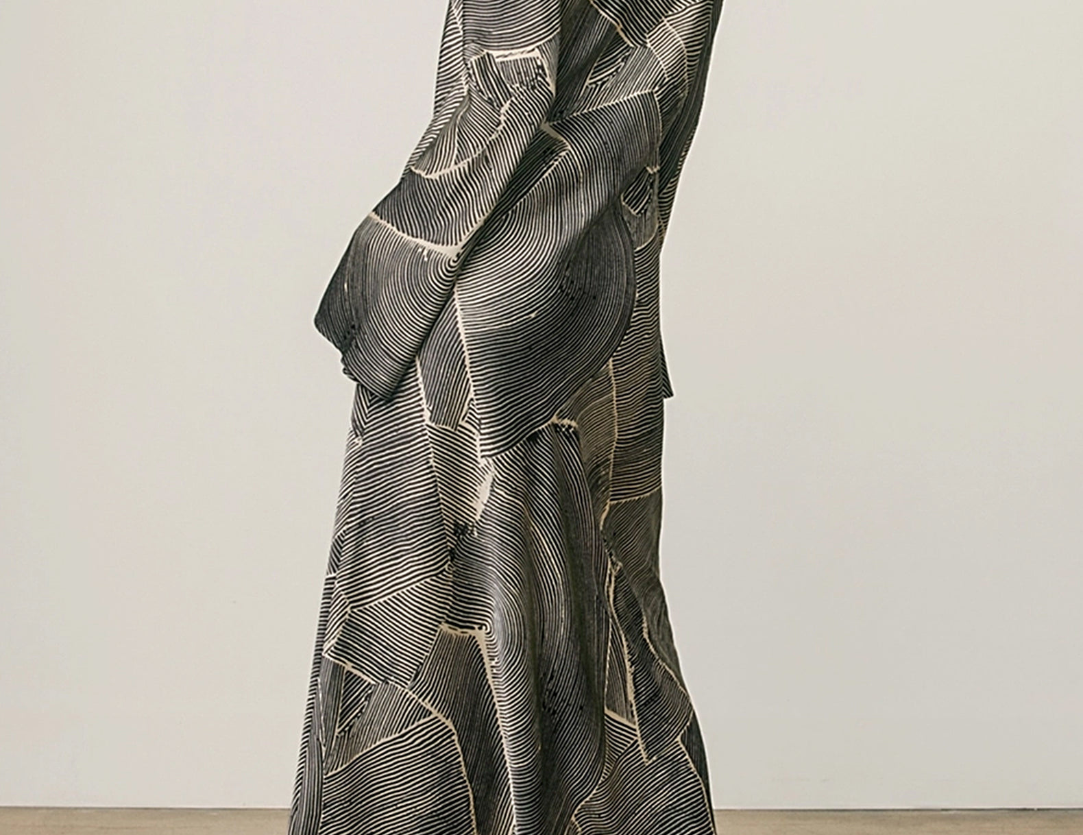A woman wearing a long-sleeved, floor-length dress with a dark gray and black abstract line pattern accented by gold geometric details, shown from a rear three-quarter angle against a neutral studio backdrop.
