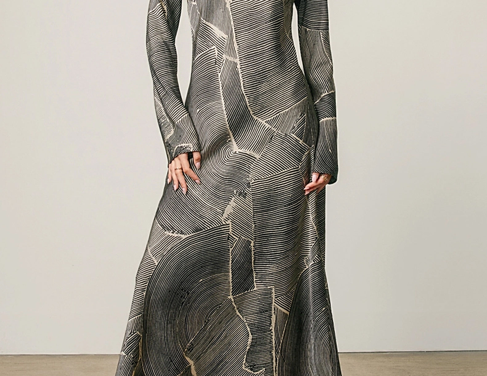 A woman modeling a long-sleeved maxi dress with a gray and black abstract wood-grain pattern accented by gold lines, paired with beige heeled sandals against a neutral studio backdrop.